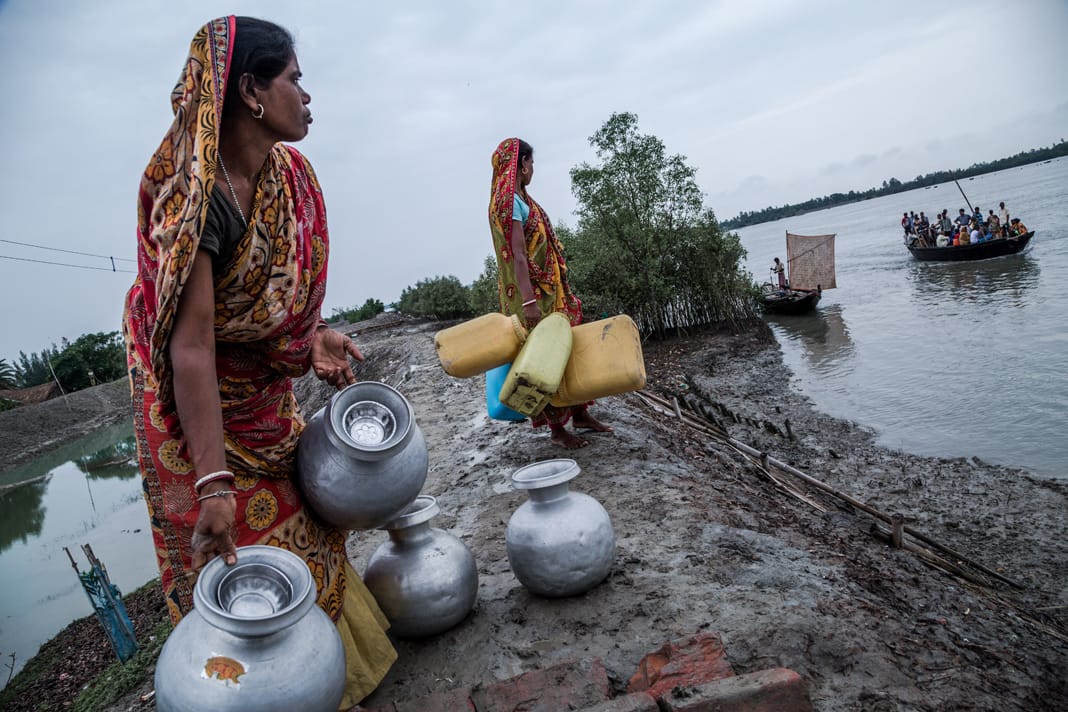 Women walk barefoot along a muddy riverbank in the Sundarbans, India, balancing metal and plastic water containers to fetch drinking water.