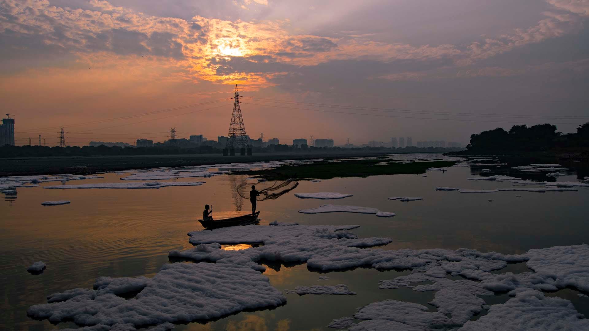 Two fishermen in a boat cast nets on the Yamuna River at sunrise, with black clouds partially covering the sun and polluted foam floating on the water.