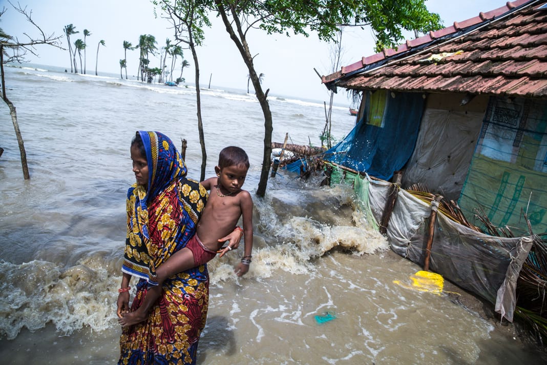 A mother holds her young child outside a flooded home on Mousuni Island, standing ankle-deep in rising seawater.