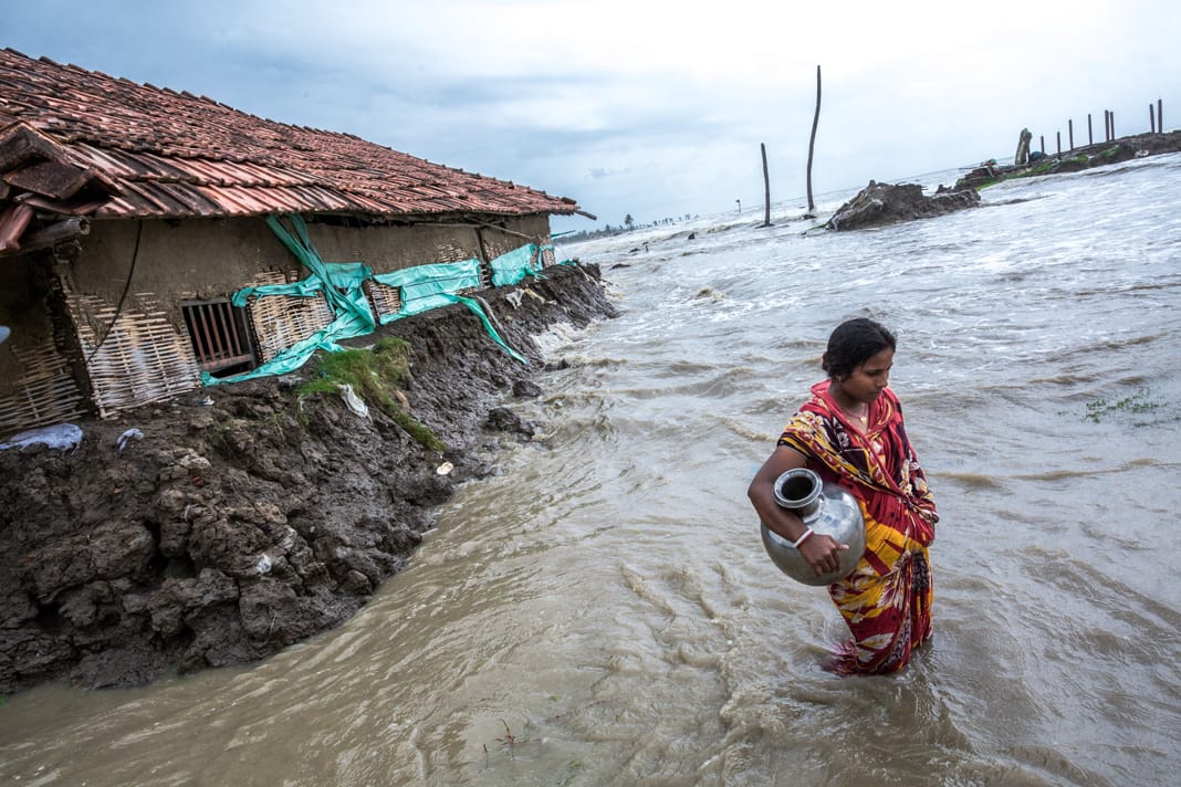 A woman wades through knee-deep floodwater carrying a metal water pot near her damaged home in the Sundarbans, India.