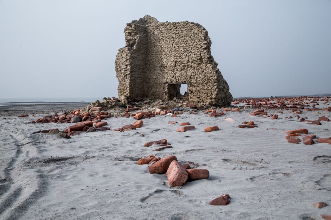 The crumbling mud walls of an abandoned house stand alone on a deserted Sundarbans shoreline, surrounded by scattered bricks.