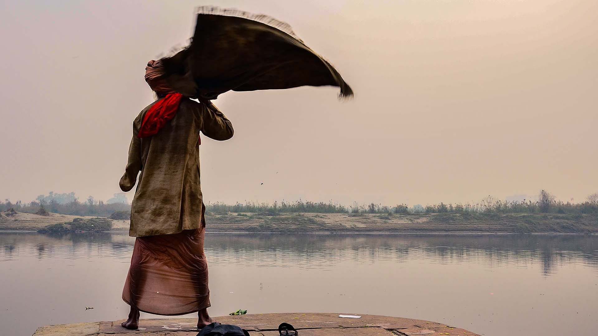 A man at Nigam Bodh Ghat lifts his cloth in the morning light, preparing for a ritual bath in the Yamuna River, where devotees worship the sun and offer lamps.