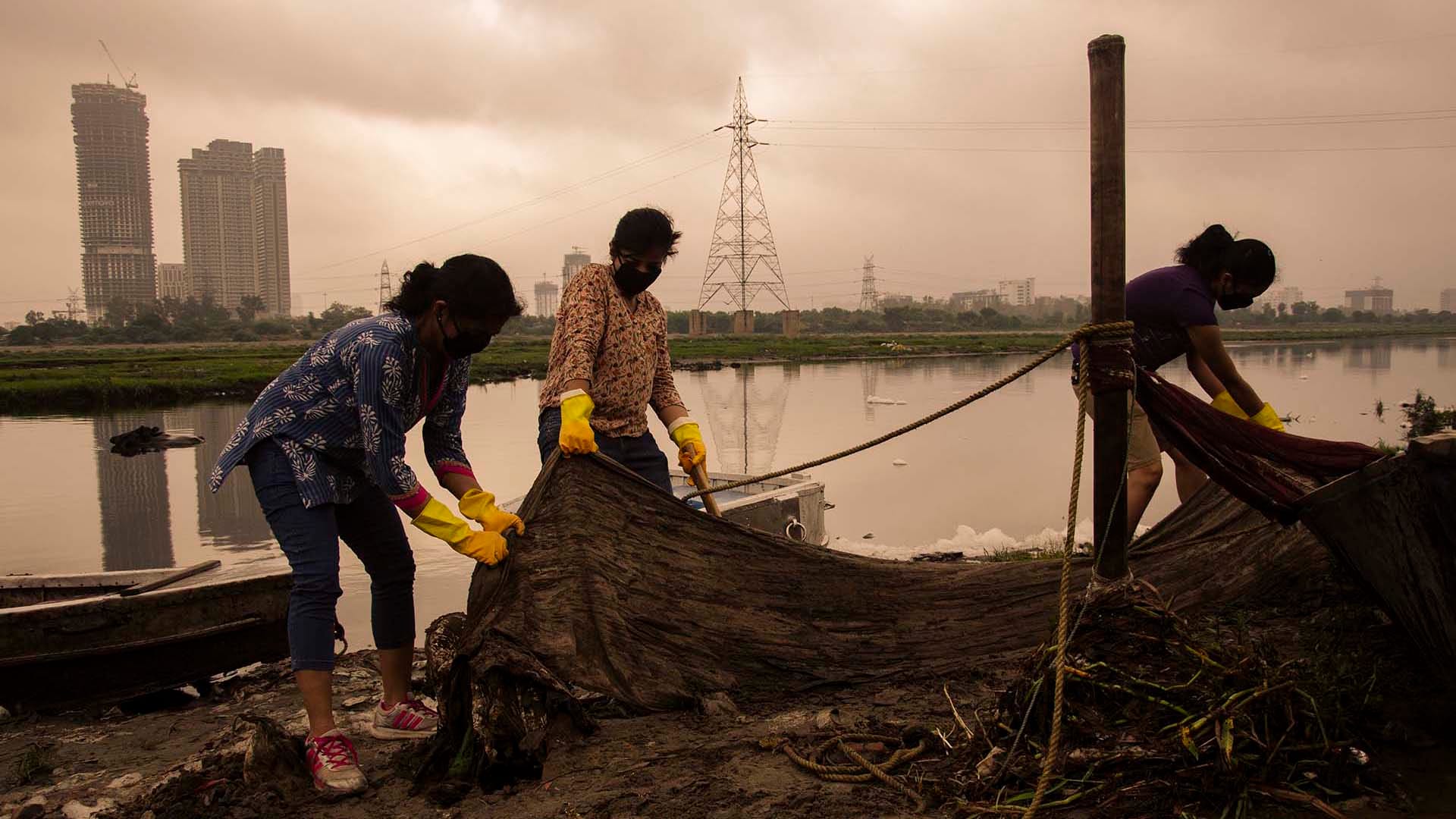 Volunteers from Namami Gange and Tree Craze Foundation remove waste from the Yamuna River, with city buildings and polluted water in the background.