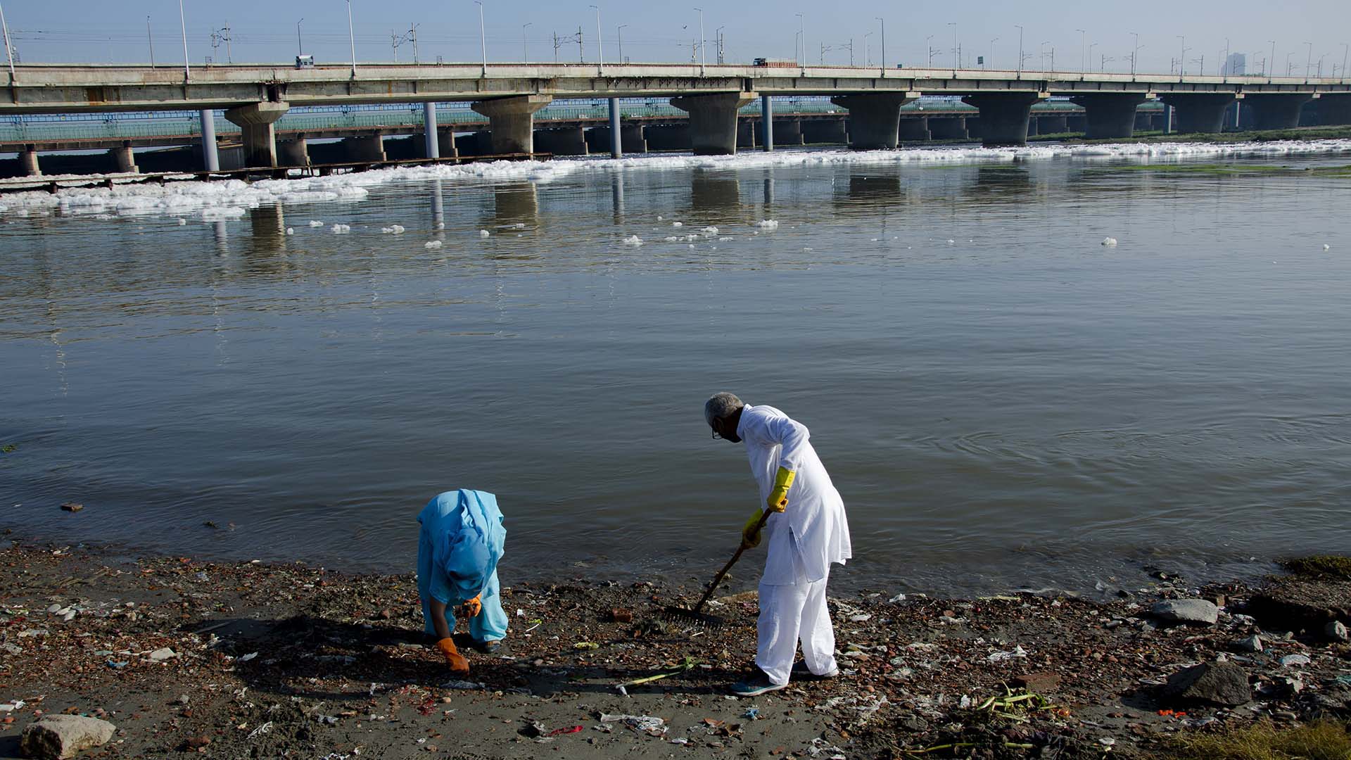 Two people clean trash from the Yamuna River’s polluted banks in Delhi, with foam patches floating on the water and a large bridge in the background.