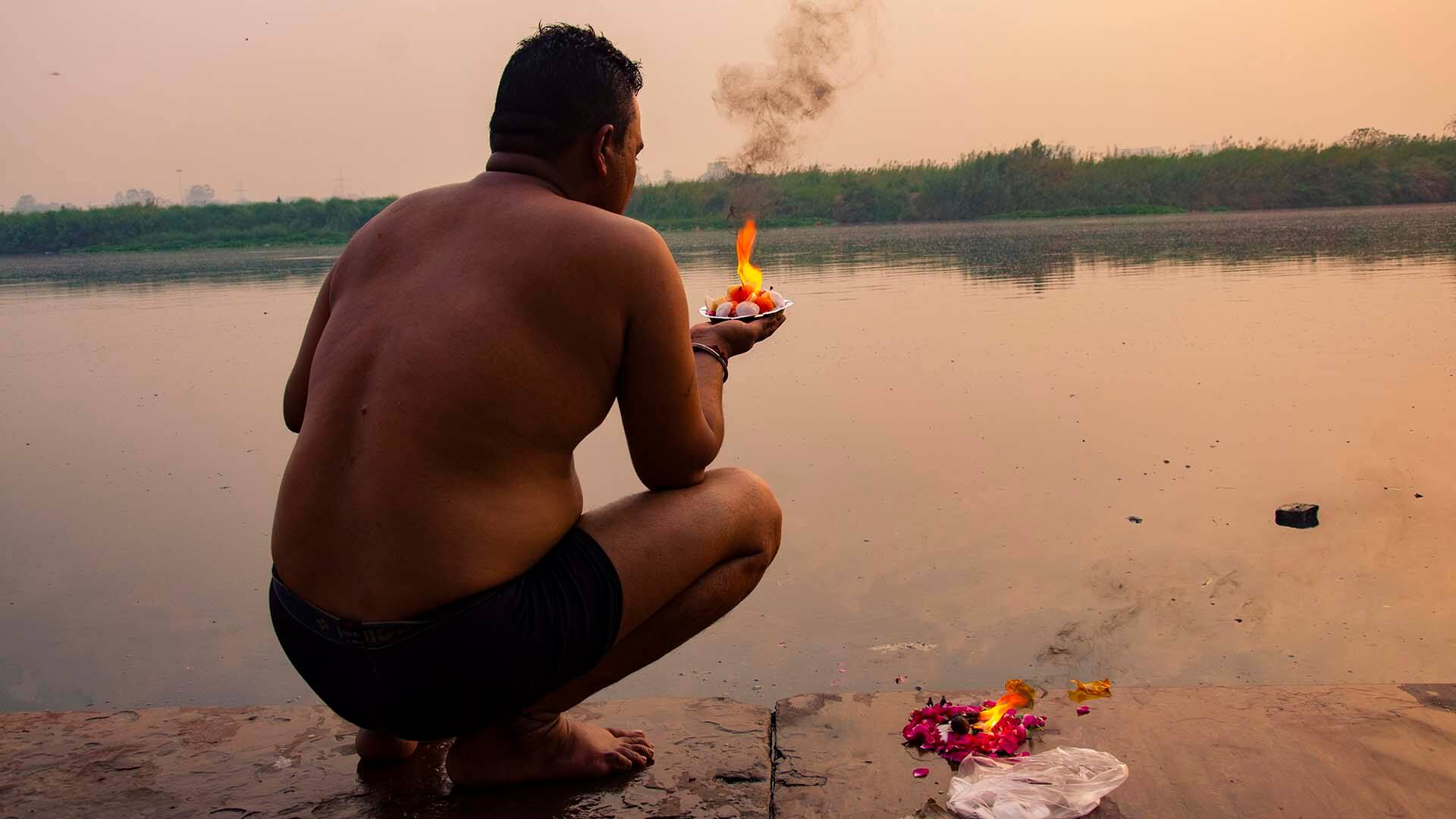 A man at Nigam Bodh Ghat kneels by the Yamuna River at sunrise, holding a lit diya (lamp) as part of his morning prayers to the sun god, Lord Surya.