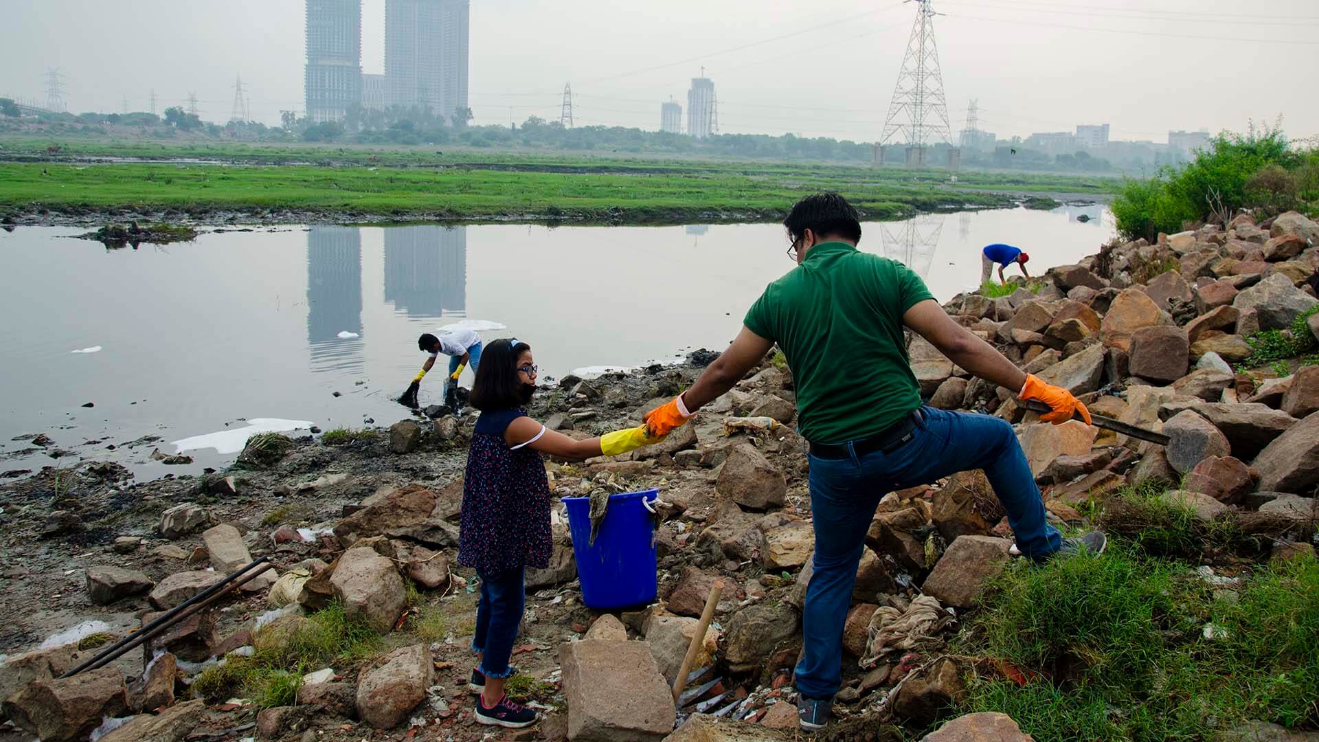 A man and a young girl wearing gloves work together to clean trash from the Yamuna River’s rocky bank, while other volunteers remove waste along the polluted river.